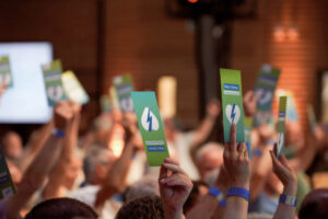Eine Gruppe von Personen hebt grüne Karten mit einem weißen Blitzsymbol in die Luft, um aktive Partizipation zu zeigen - vielleicht bei einer Veranstaltung der Elektrizitätswerke Schönau (EWS). Der Fokus liegt auf den erhobenen Karten und Händen, der Hintergrund ist unscharf.