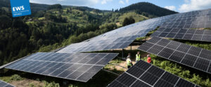 Auf einem grasbewachsenen Hügel sind Reihen großer Solarmodule installiert, die von vier Personen in Sicherheitswesten inspiziert werden. Im Hintergrund sind bewaldete Hügel und ein paar Häuser zu sehen, der Himmel ist teilweise bewölkt.