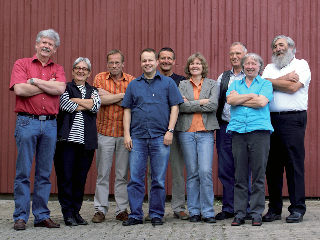 Eine Gruppe von neun Bürgerinnen und Bürgern steht draußen vor einer roten Holzwand, lächelt und posiert mit verschränkten Armen oder Händen an der Seite - sie repräsentieren den Geist des Stromwechsels bei den Elektrizitätswerken Schönau.