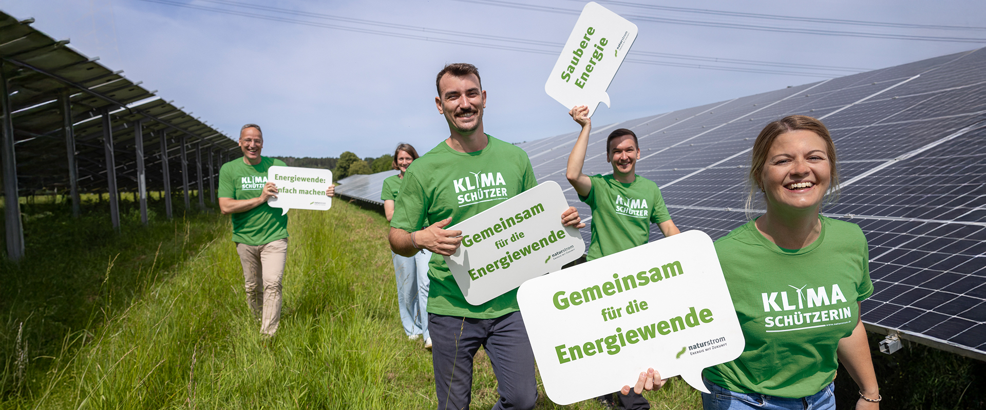 Vier lächelnde Menschen stehen neben den Solarpark-Paneelen, halten Schilder mit der Aufschrift Gemeinsam für die Energiewende und tragen grüne KLIMASCHÜTZER:IN-Shirts. Es ist ein sonniger Tag mit klarem Himmel, perfekt für die Ökostromerzeugung.