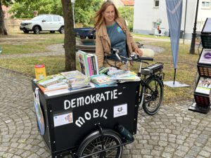Eine Frau steht im Freien neben einem Lastenfahrrad mit der Aufschrift „DEMOKRATIE-MOBIL“, das mit Büchern und Broschüren beladen ist. Die Szene spielt in einer parkähnlichen Umgebung, in der Bäume, Kopfsteinpflaster und geparkte Autos zu sehen sind.