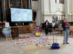Zwei Frauen stehen in einem Kirchenraum, auf dem Boden liegt eine bunte Patchworkdecke, auf einer Leinwand werden eine Karte und Fotos gezeigt, und es hängen Banner von „Omas for Future“. Eine Frau spricht, während die andere neben dem Podium steht.