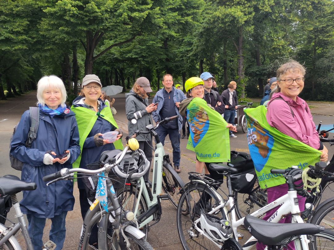 Eine Gruppe von Menschen, überwiegend ältere Erwachsene, steht mit Fahrrädern in einem Park. Vier Frauen im Vordergrund lächeln und tragen grüne Umhänge oder Westen. Im Hintergrund sind unter bewölktem Himmel Bäume und weitere Teilnehmer zu sehen.