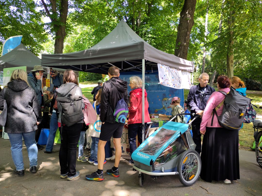 In einem grünen Park versammelt sich eine Gruppe von Menschen, darunter Erwachsene und Kinder, um einen Stand mit einem grauen Sonnensegel. Einige Erwachsene tragen Rucksäcke, und es sind Kinderwagen zu sehen. Im Hintergrund sind Bäume und Sonnenlicht zu erkennen.