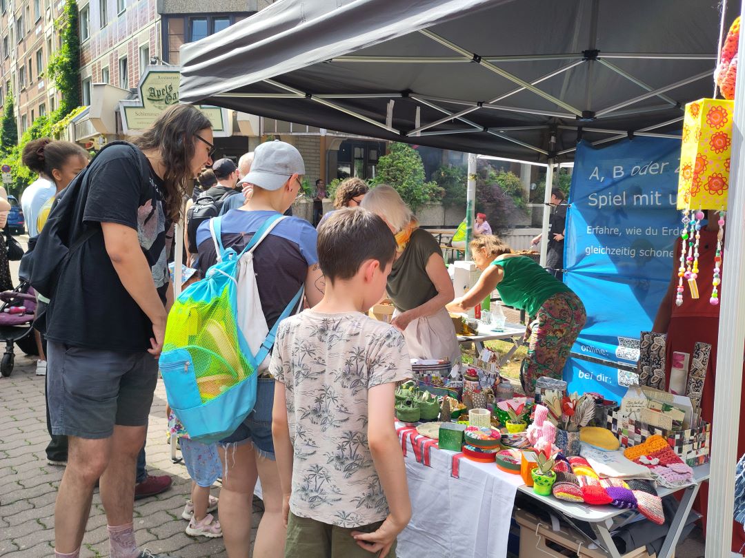 Eine Gruppe von Menschen, darunter Kinder und Erwachsene, begutachtet handgefertigte Kunsthandwerksartikel und bunte Waren, die auf Tischen unter dem Vordach eines Marktstandes auf einem Straßenfest im Freien ausgestellt sind. Im Hintergrund ist ein blaues Informationsbanner zu sehen.