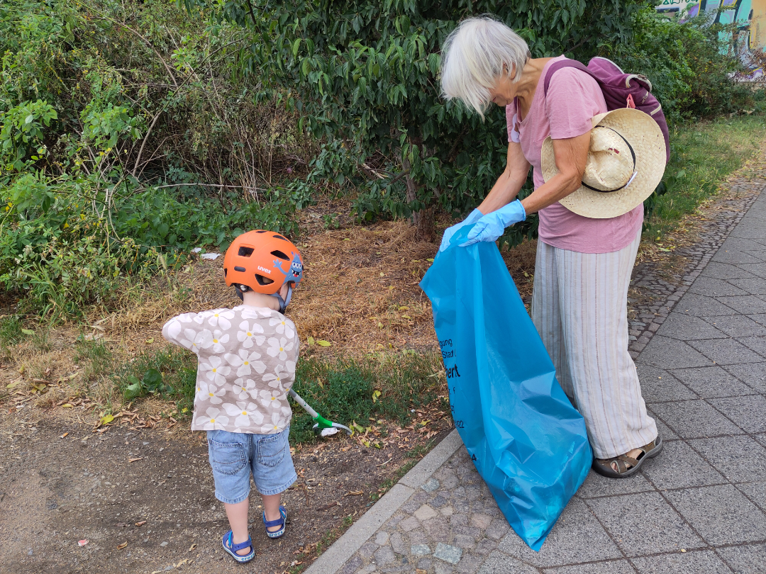Eine ältere Frau und ein kleines Kind, das einen Helm trägt, sammeln gemeinsam neben einem Gehweg Müll ein. Die Frau hält einen großen blauen Müllsack und einen Strohhut in der Hand, während das Kind mit einer Greifzange den Müll aufsammelt.