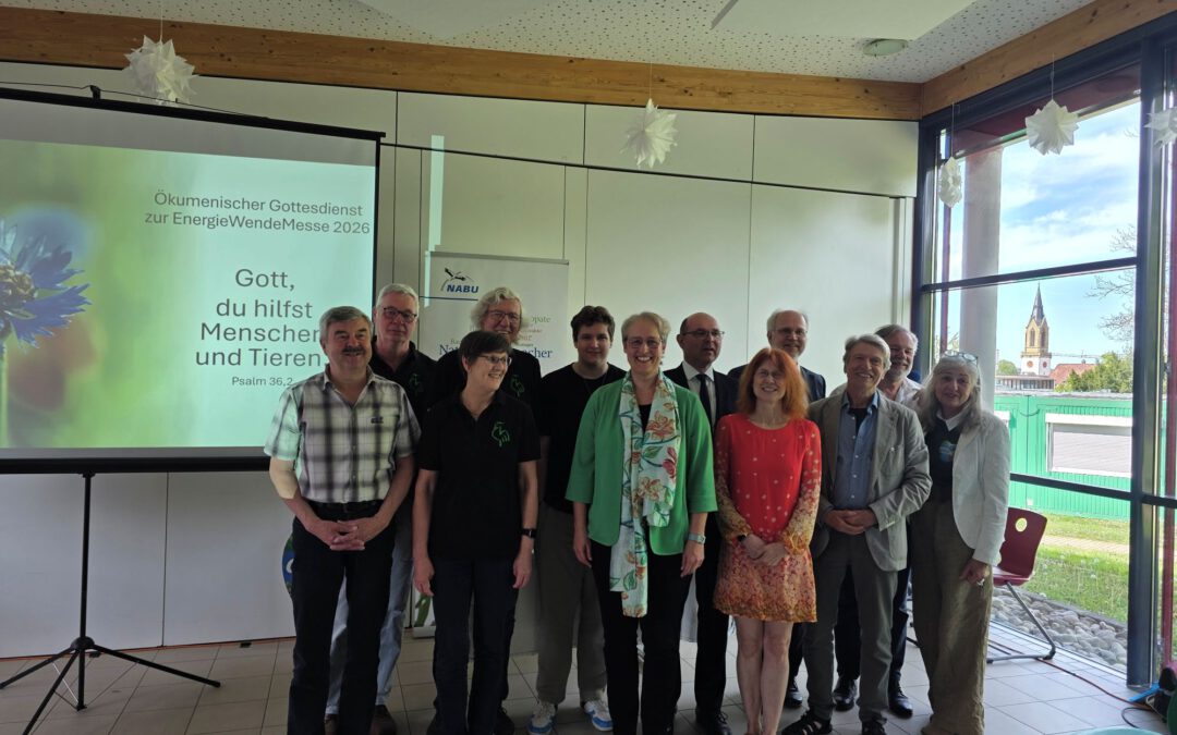 Eine Gruppe von 13 Erwachsenen steht lächelnd in einem Raum mit großen Fenstern. Hinter ihnen ist auf einer Leinwand ein deutscher Text über einen Gottesdienst und ein Psalmvers zu sehen. Durch die Fenster sind eine Landschaft und eine Kirche zu erkennen.