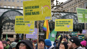 Eine Menschenmenge bei einem Klimaprotest hält Schilder auf Deutsch und Englisch, darunter GUTES KLIMA GIBT'S NUR OHNE NAZIS und ALL YOU NEED IS LOVE AND ZUKUNFTSFÄHIGE KLIMAPOLITIK. Die Menschen sind in Winterkleidung eingepackt.