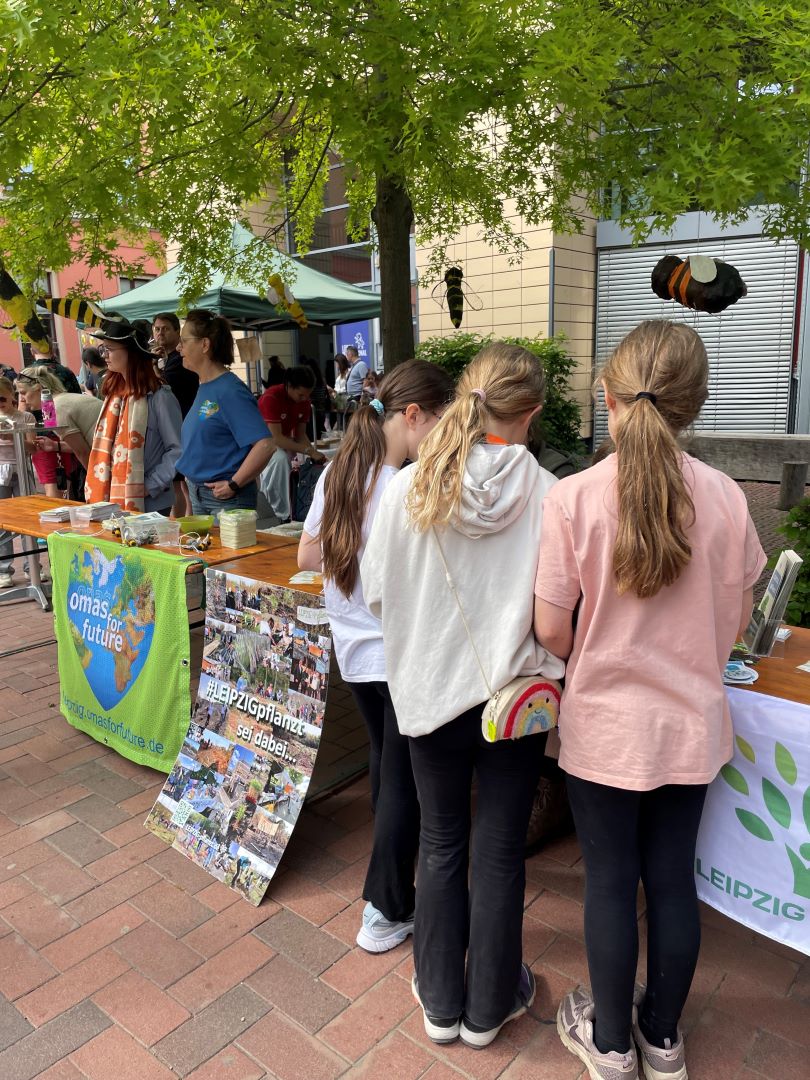 Drei Mädchen stehen an einem Informationsstand im Freien mit grünen Bannern, auf einem davon steht „Omas for Future“. Am Stand liegen Broschüren und Fotos aus, im Hintergrund sind Menschen und Bäume zu sehen.