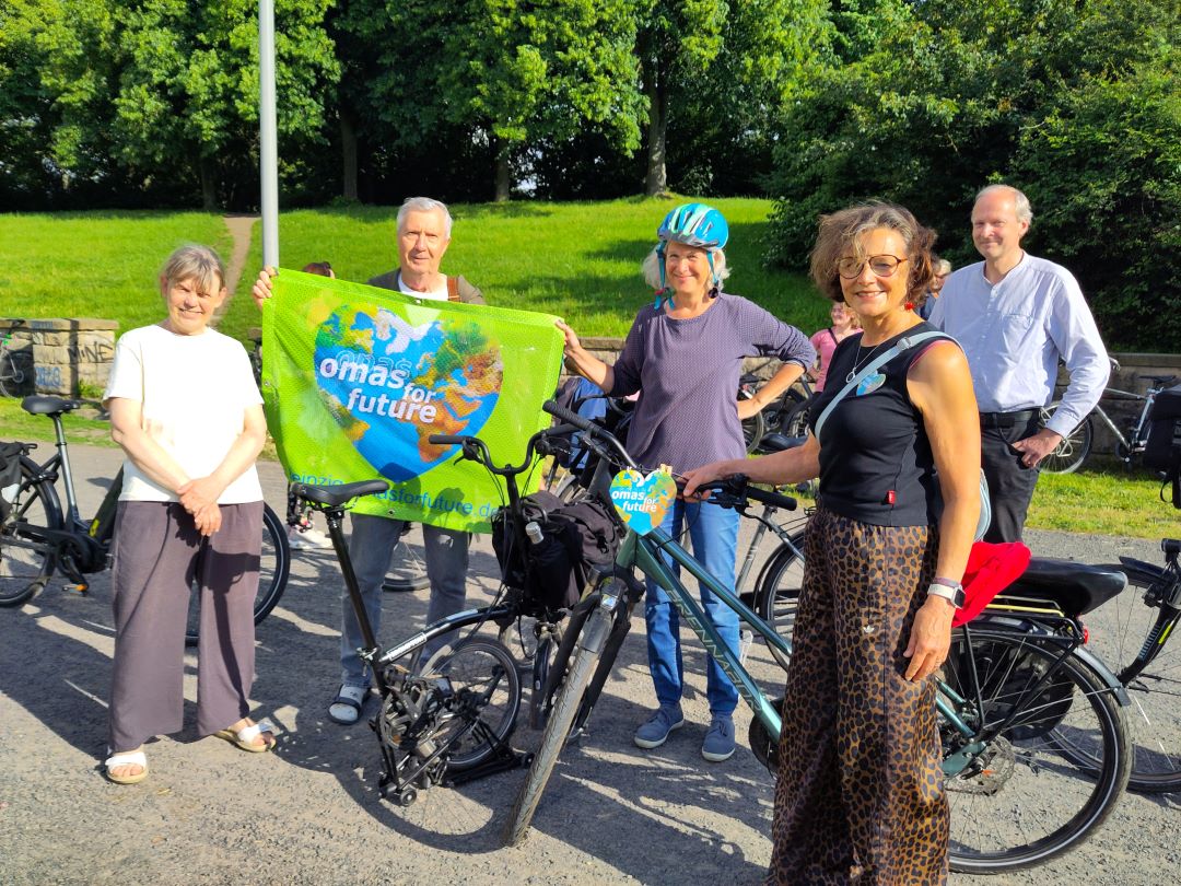 Fünf Erwachsene stehen mit Fahrrädern im Freien und lächeln in die Kamera. Zwei von ihnen halten ein grünes Transparent mit der Aufschrift „Omas for Future“ hoch. Im Hintergrund sind Bäume und Sonnenlicht zu sehen, was eine fröhliche Atmosphäre schafft.