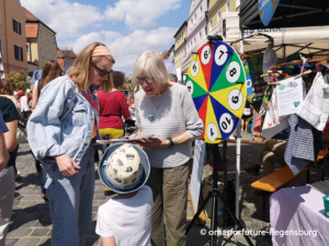 Zwei Frauen und ein Kind stehen auf einem Straßenfest im Freien neben einem bunten Spinnrad. Eine Frau spricht mit dem Kind, während sie ein Klemmbrett in der Hand hält. Stände, Menschen und Gebäude säumen die Kopfsteinpflasterstraße.