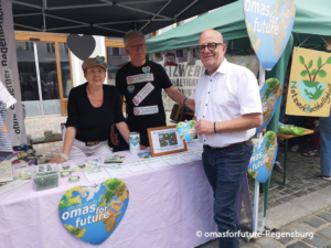 Drei Personen stehen an einem Stand im Freien, der mit Schildern mit der Aufschrift „Omas for Future“ geschmückt ist, die für Nachhaltigkeit werben. Am Stand liegen Flyer, Ansteckbuttons und eine Spendendose aus. Zwei Personen stehen hinter dem Tisch, eine weitere Person steht davor und hält ein herzförmiges Schild in der Hand.