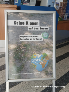 Auf einem deutschen Schild steht „Keine Kippen auf den Boden!“, zusammen mit einem Hinweis auf kostenlose Aschenbecher an der Theke und einem „Omas for Future“-Logo in Form eines Herzens mit einer Weltkarte. Das Schild steht auf einem Gehweg im Innenbereich.