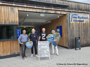 Vier ältere Menschen stehen vor dem Stadtwerk Wöhrdbad, halten herzförmige Schilder mit der Aufschrift „Omas for Future“ in den Händen und lächeln. Vor ihnen steht auf einer Staffelei ein Plakat zum Thema Klimaschutz.