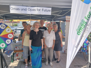 Eine Gruppe älterer Menschen steht lächelnd unter einem Zelt, an dem ein Banner mit der Aufschrift „Omas und Opas für die Zukunft“ hängt. Auf den Tischen um sie herum liegen bei einer Veranstaltung im Freien ein buntes Glücksrad und Informationsmaterialien.