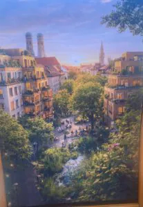Eine lebendige Stadtlandschaft bei Sonnenuntergang zeigt üppiges Grün, Wohnhäuser mit Balkonen, Menschen, die sich auf einem Platz versammeln, und zwei Kirchtürme im Hintergrund unter einem bunten Himmel.