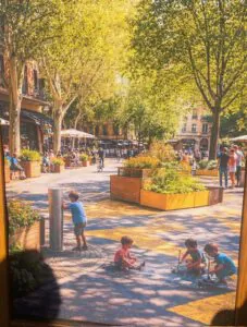 Ein sonnenbeschienener Stadtplatz mit Menschen, die in Straßencafés sitzen, grüne Bäume säumen den Platz. Im Vordergrund spielen Kinder auf dem bemalten Pflaster, ein Kind an einem Wasserbrunnen, während andere mit Kreide zeichnen.